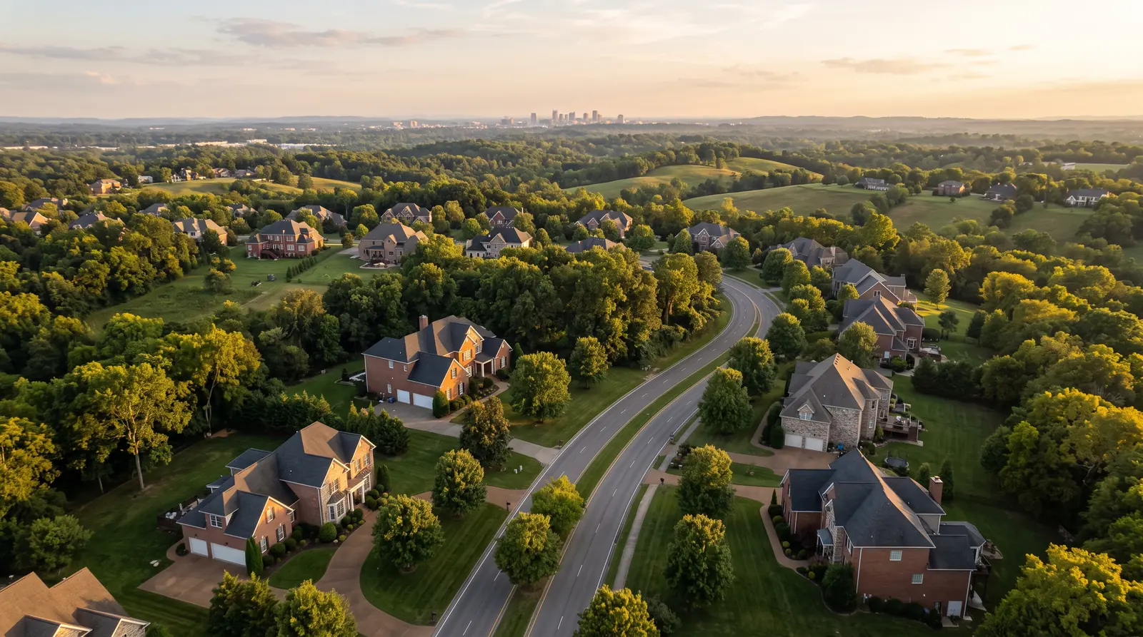 An aerial view over a green Middle Tennessee neighborhood at golden hour showing brick and stone homes, mature trees and rolling hills, a winding road and a glimpse of a city skyline far away