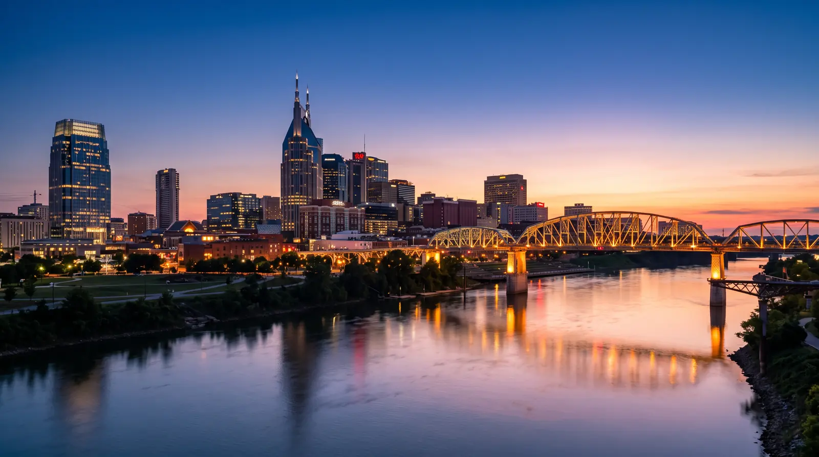 Nashville Skyline at Blue Hour | Music City Cleaner The Nashville skyline glowing at blue-hour dusk beyond the Cumberland River with a calm reflection, a warm sunset gradient in the sky and the lit pedestrian bridge
