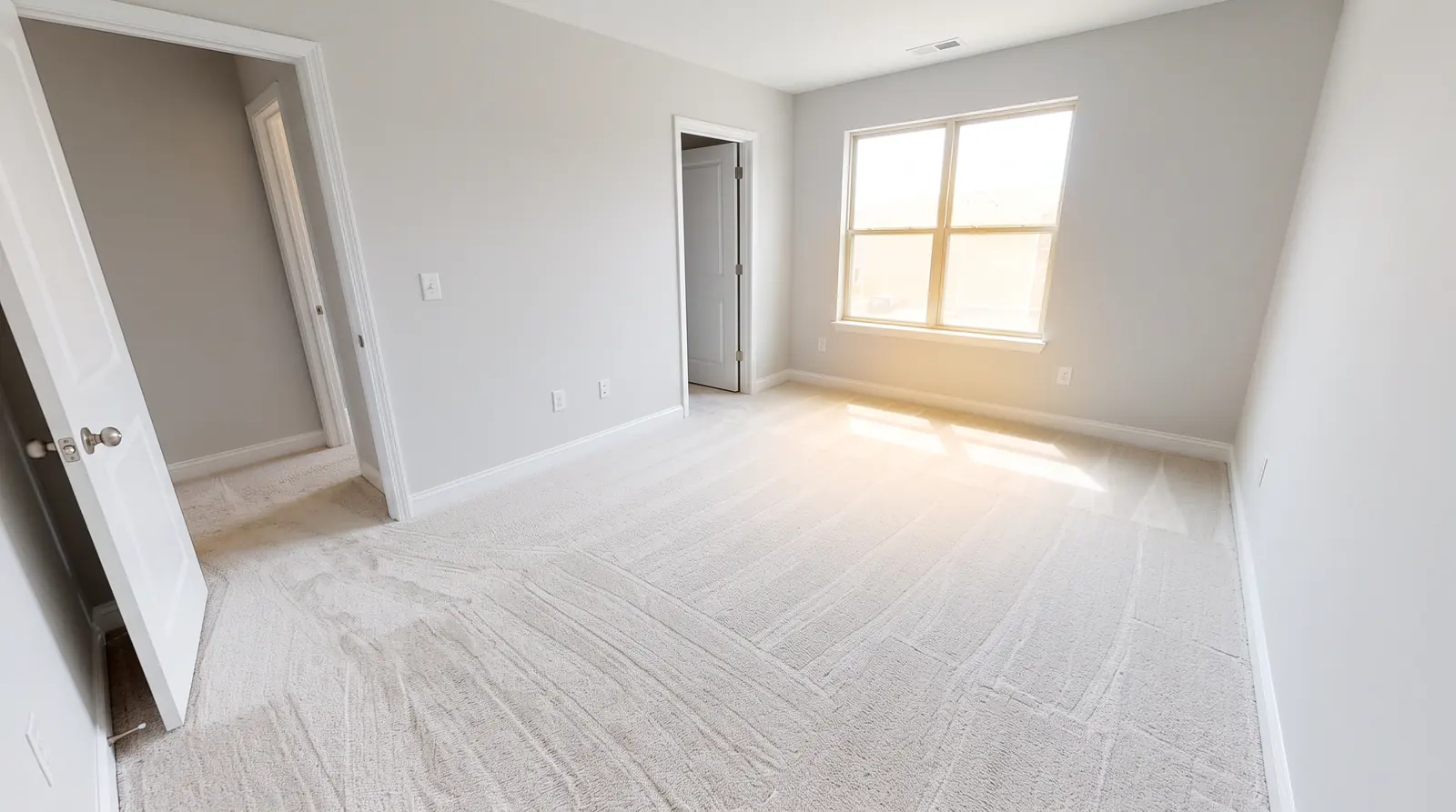 A freshly cleaned empty apartment bedroom with vacuum lines on pale carpet, spotless window and baseboards, the door ajar and sunlight on the floor
